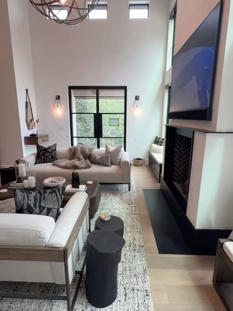 Installed and styled living room of a Lake Austin residence — double-height ceiling, chandelier, fireplace with herringbone tile and wall-mounted TV, steel-frame glass doors